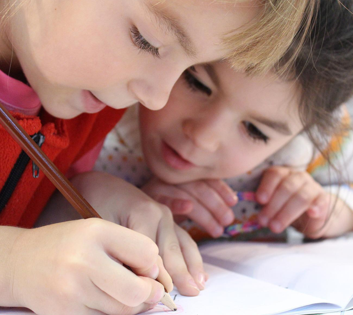 Two children with their heads close together writing and looking closely at a notebook.