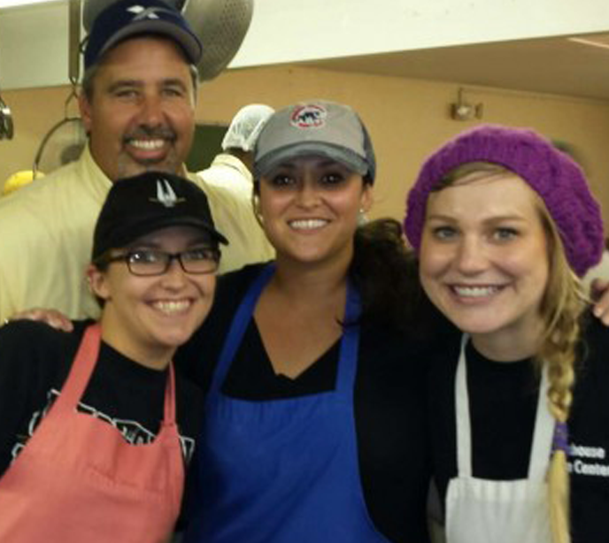 A group of three women and one man stood together wearing hats and aprons smiling in a kitchen.