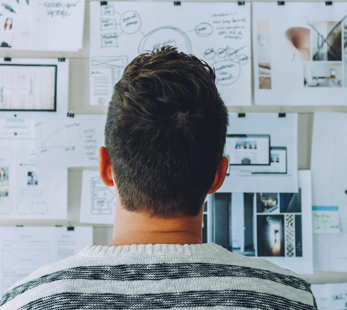 The back of a man's head wearing a striped sweater looking at multiple papers on a drawing board.