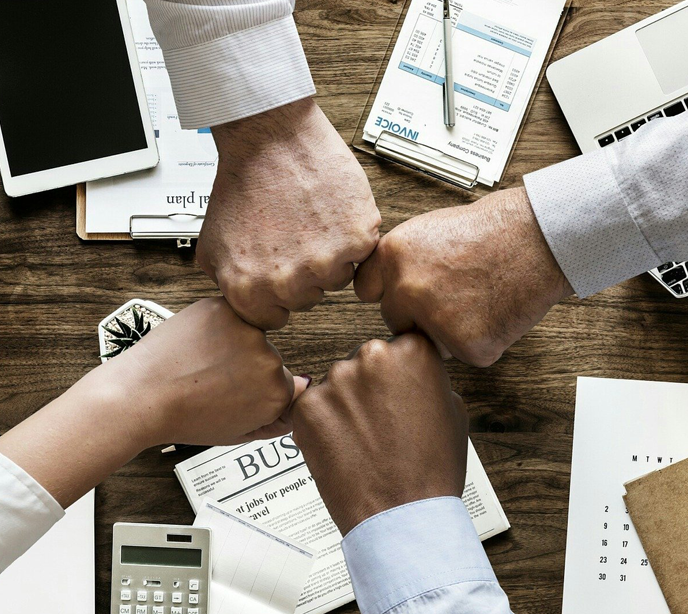 Four arms stretched out with fists touching in a cross over a table with clipboards with invoices, tablets and calculators.