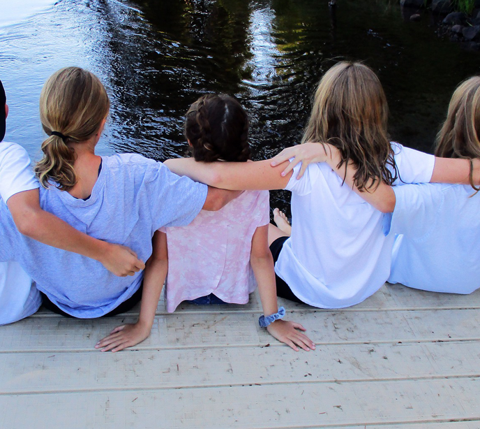 Five Children sitting on a dock wearing pale coloured t-shirts linking arms over their shoulders