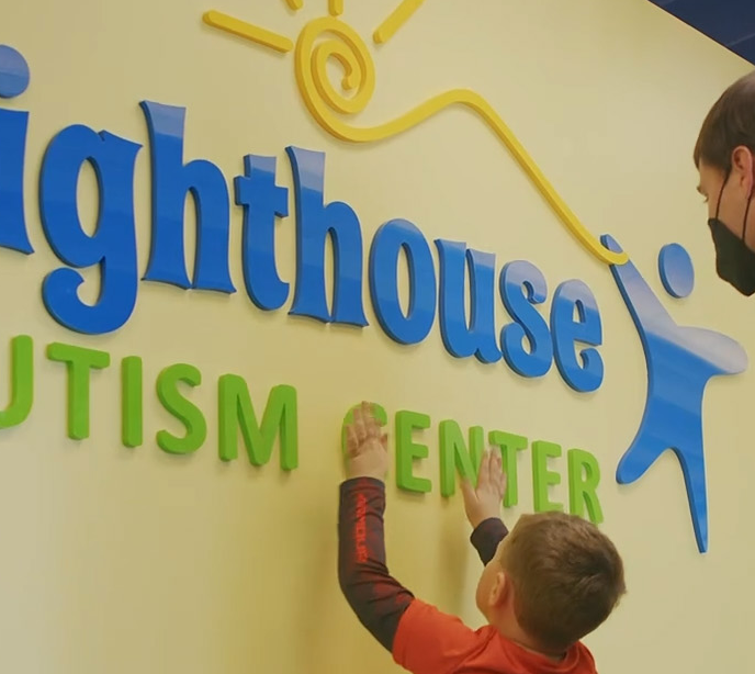 A child wearing an orange shirt with both of his hands on a yellow wall touching a Lighthouse Autism Center logo.