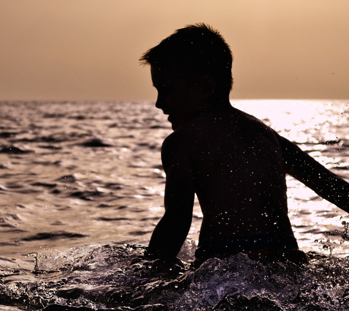 A silhoutte of a child stood in a low tide of the ocean splashing water around him at dusk.