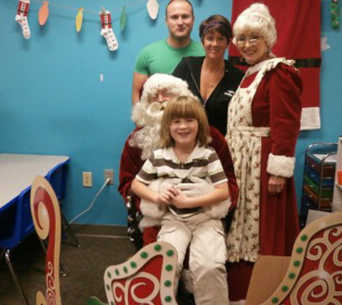 Santa Claus with a child sat on his knee and his wife stood with the child's parents all in front of a cardboard sleigh and holiday decor.