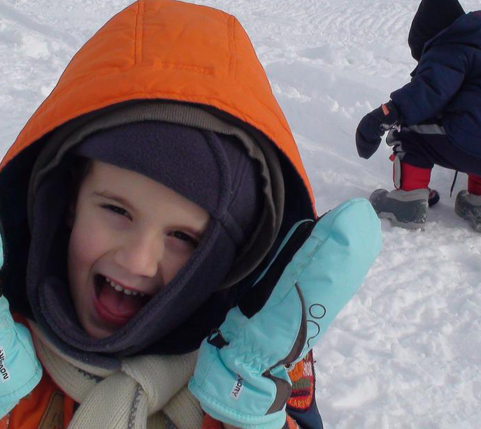 A child wearing multiple layers of warm clothing stood in the snow smiling and a child crouched down picking up snow.