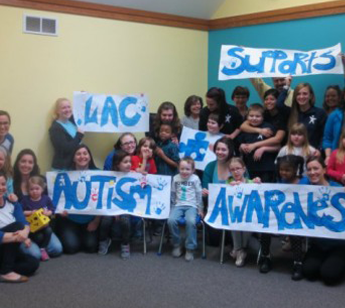 Lighthouse Autism Center staff members and children sat in a semi-circle holding LAC Supports Autism Awareness blue banners.