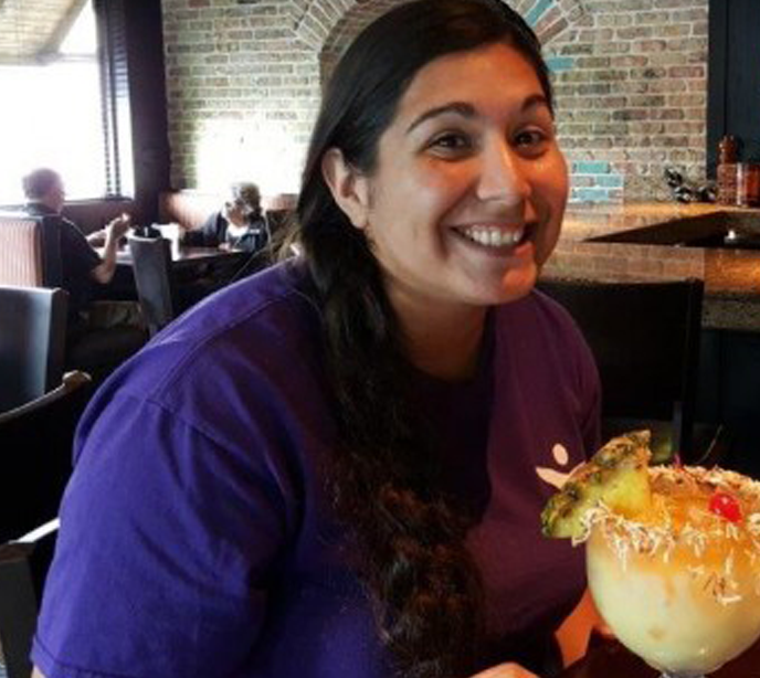 A Lighthouse Autism Center employee wearing a blue shirt smiling at a table with a piña colada cocktail.