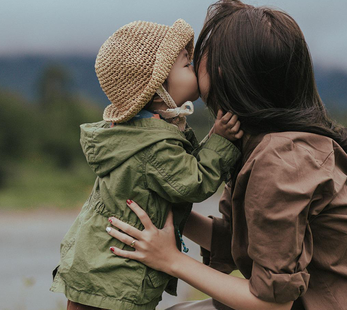 A child wearing a brown woven hat and green coat giving his Mom a kiss on the cheek