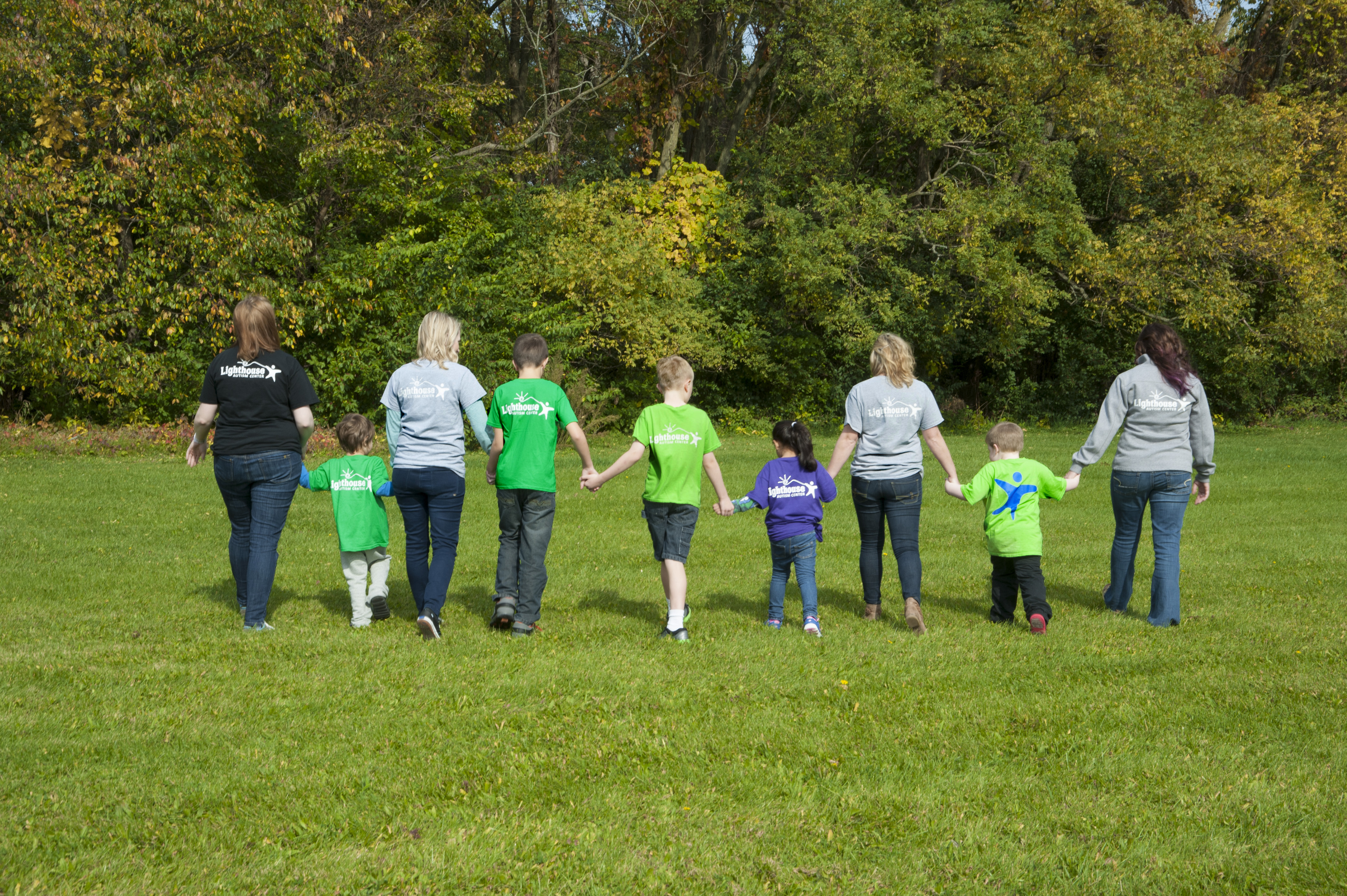 Four Lighthouse Autism Center staff members holding hands in a line with children in branded t-shirts on the grass facing trees