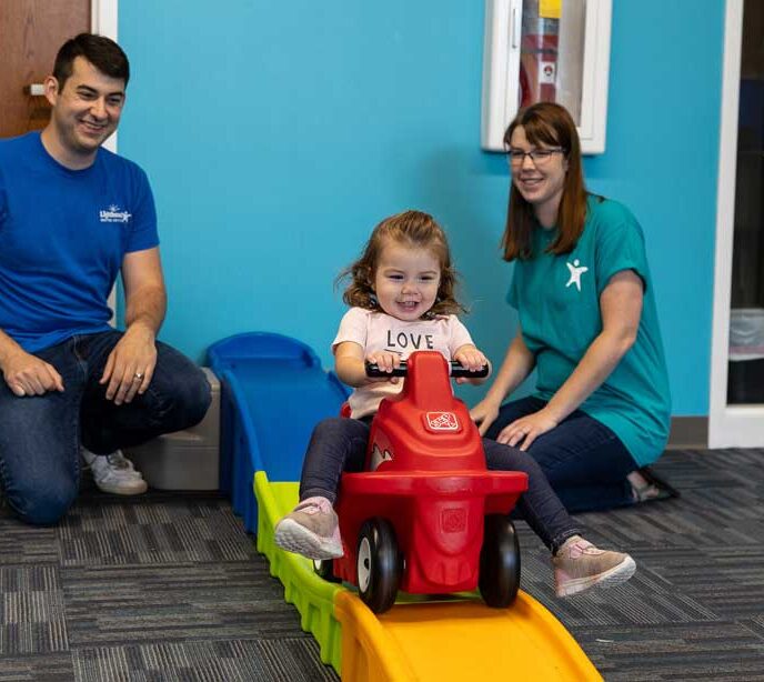 Two Lighthouse Autism Center employees smiling at a child on a toy motorbike going down a slide