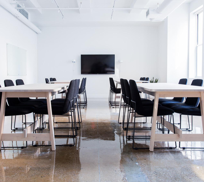 A boardroom with four white tables and black chairs with white walls and floors in front of a television on the wall