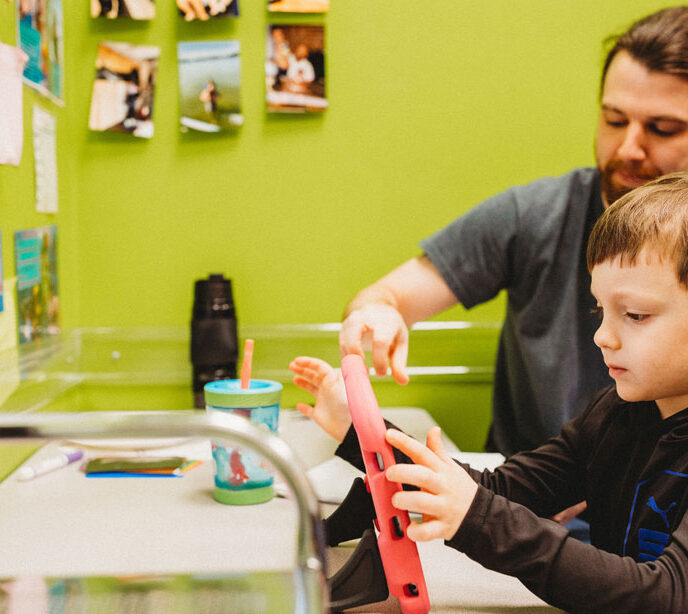 A child in a green room playing on a tablet with a Lighthouse Autism Center member of staff