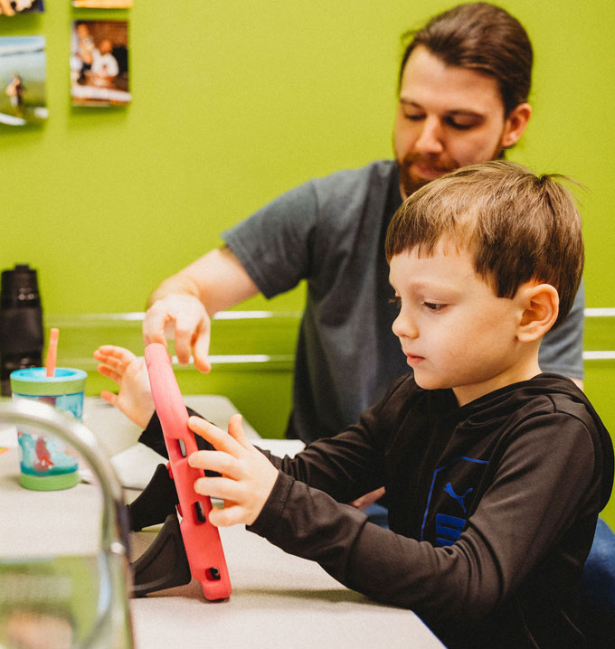 A young boy holding a tablet sat at a table with a Lighthouse Autism Center staff member helping him