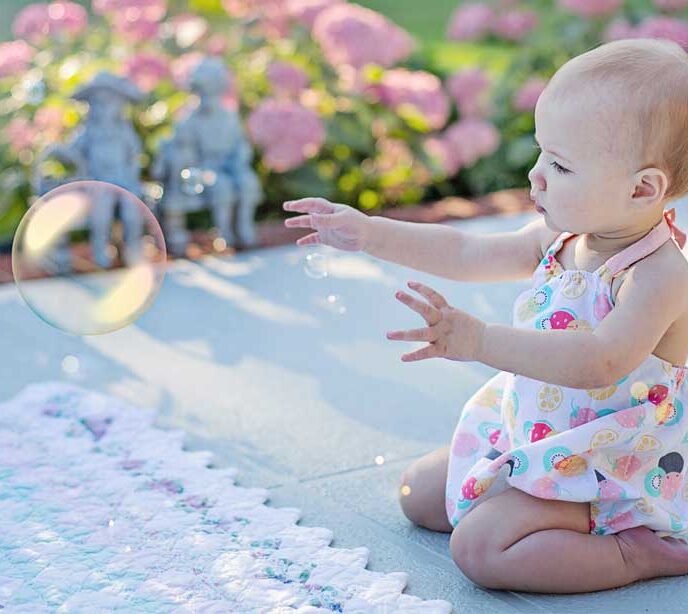 A baby wearing a white dress with a fruit pattern knelt down on a blanket and their arms stretched out to bubbles