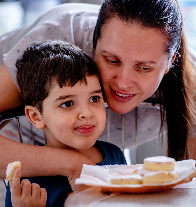 A woman with a pony tail with her arm around a young boy eating some dessert from a plate.