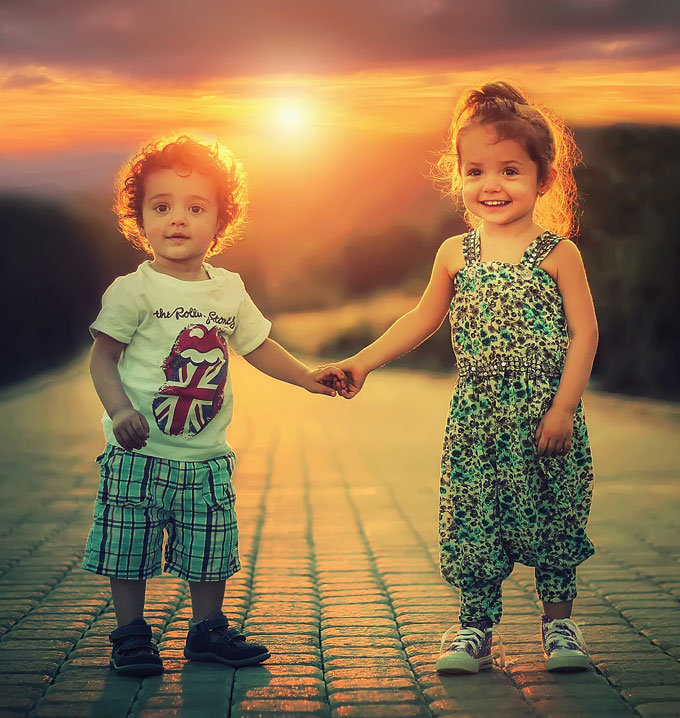 A young boy wearing blue shorts and a white shirt holding hands with his sister in a blue outfit in front of a sunset