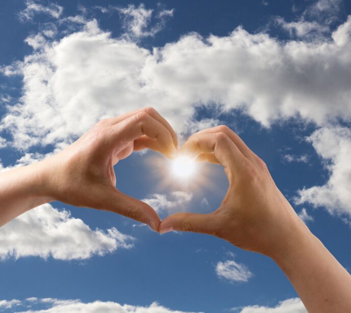 A pair of hands making a heart symbol in front of a blue sky with clouds and the sun shining in the center