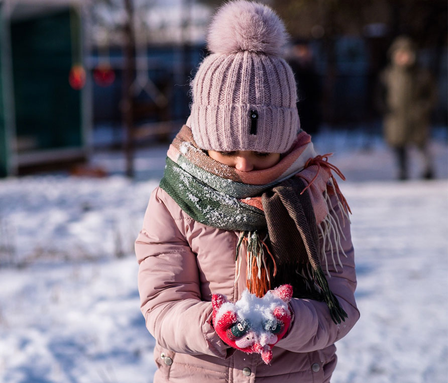 A child wearing a pink hat, jacket, scarf and gloves holding a small ball of snow in her hands.