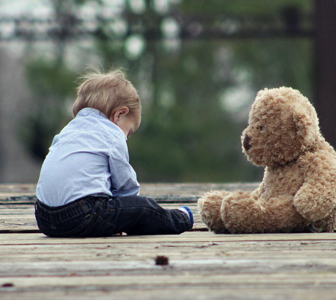 A child sat on a decking wearing a blue shirt and jeans hunched over next to a teddy bear.