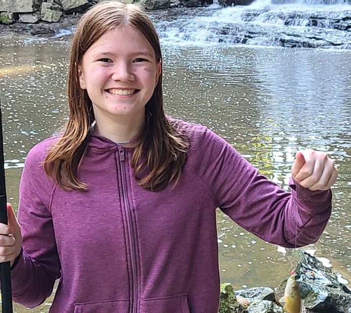 A girl called Ali stood in front of a lake smiling holding a fish on a piece of chord in one hand and a fishing pole in the other