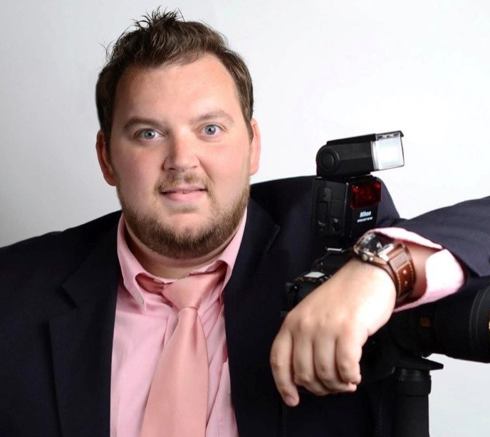 Christopher Adams wearing a suit with a pink shirt and a camera under his arm in front of a white background