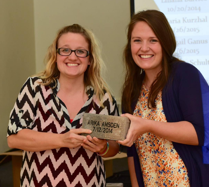 A brunette woman and a blond woman earing glasses stood together holding a brick with writing on it saying 