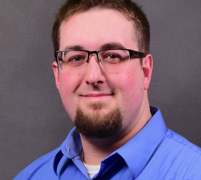 Sean Addley wearing a blue shirt smiling in front of a gray background