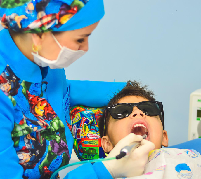 A dentist wearing a uniform with superhero print and a child in a dental chair wearing sunglasses for a checkup