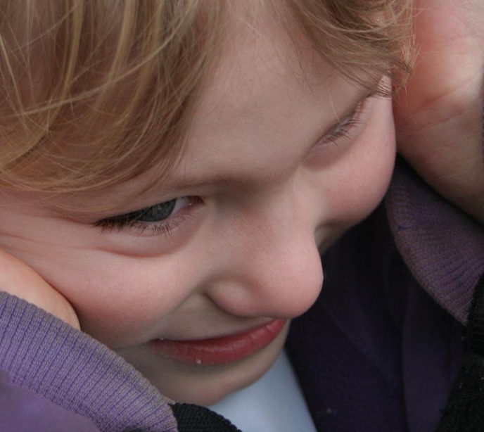 A close-up shot of a child smiling with blonde hair wearing a purple sweater with her hands over her ears