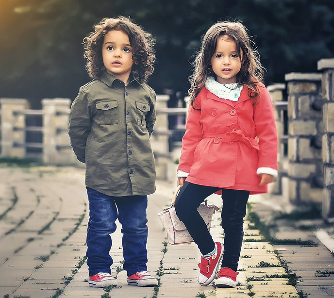 A male child wearing blue jeans and a green shirt beside a female child wearing a red coat, shoes and jeans