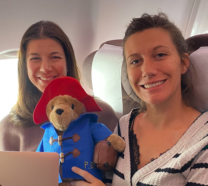 Two ladies smiling sat inside an airplane holding a Paddington bear toy
