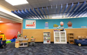 A toy play Fresh Mart shop with toy plastic cars, a toy cash register, shelving units with toy food with blue walls and carpet