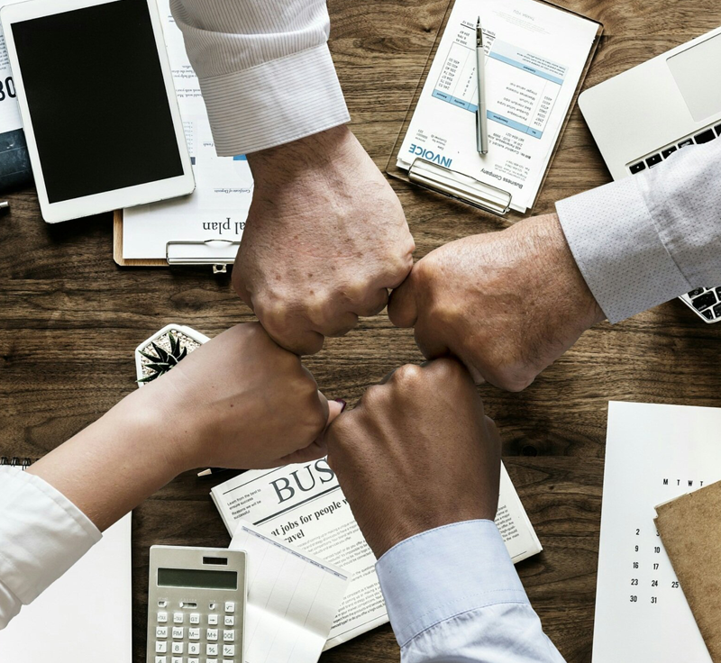 Four arms holding their hands in fists in the center above a brown table with invoices, a calculator and a laptop