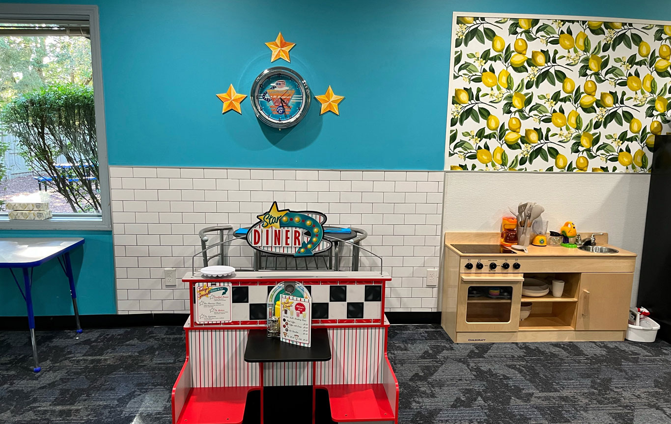 A zoomed out version of a toy wooden kitchen with lemon wallpaper above, a toy star diner and a clock on a blue wall.