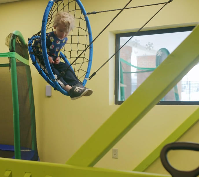 A child sat on a netted swing seat in a yellow room swinging from one side of the room to the other