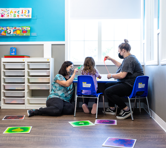 Two Lighthouse Autism Center employees sat with a child at a table wearing face masks playing with colored putty