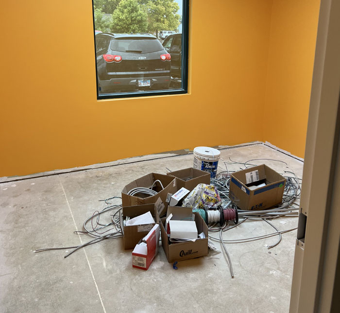A room with yellow walls and a window in the center and cardboard boxes, electrical wires and paint pots on the floor