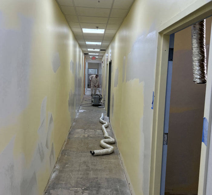 A hallway with pale yellow walls and a person wearing a white decorator's suit repainting the wall