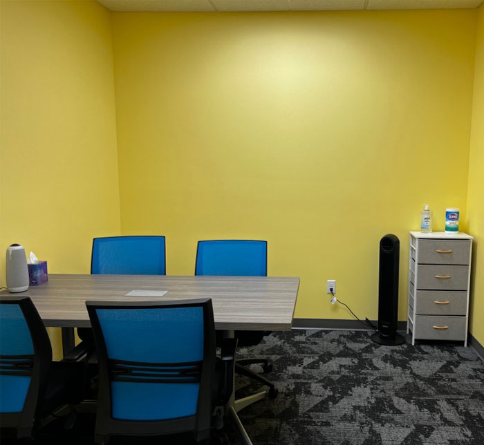 A conference room with yellow walls, a gray table and four blue desk chairs at Lighthouse Autism Center