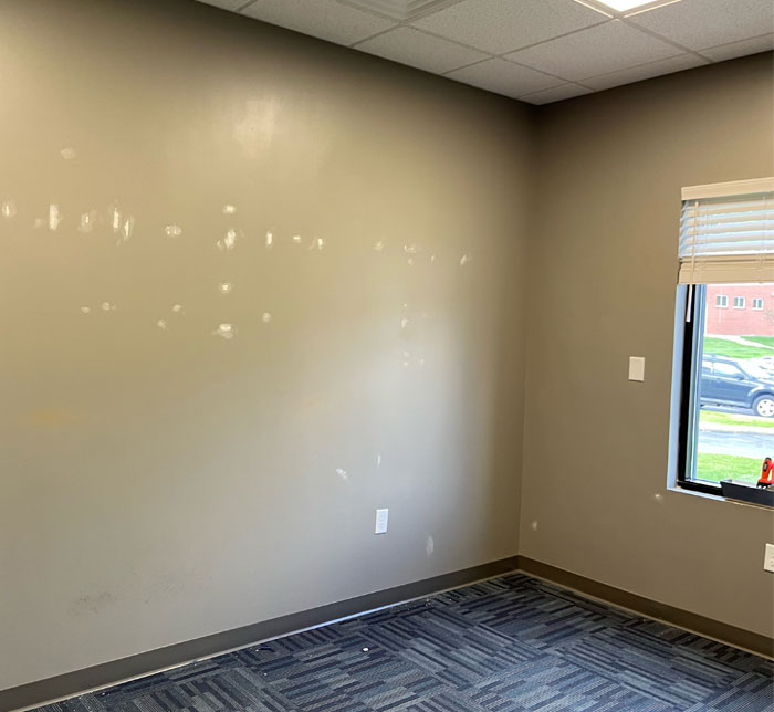 A therapy room under construction with a beige wall and spots of white paint with a blue carpet and window with the blinds open at South Bend Autism Center