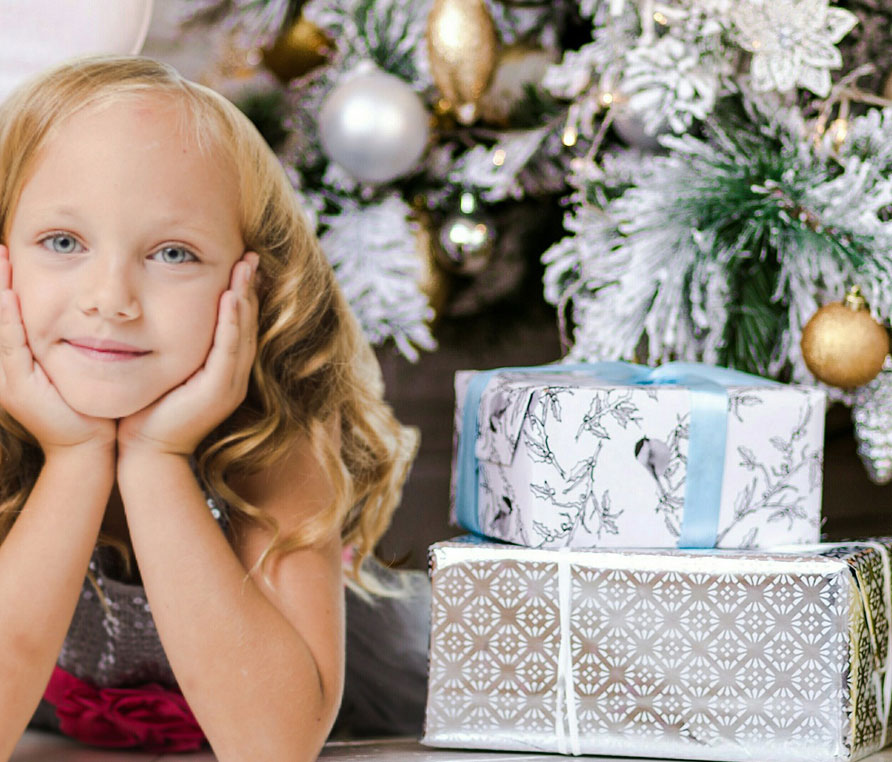 A child with blonde hair and her hands covering her ears laying next two silver gifts and a white and gold christmas tree.