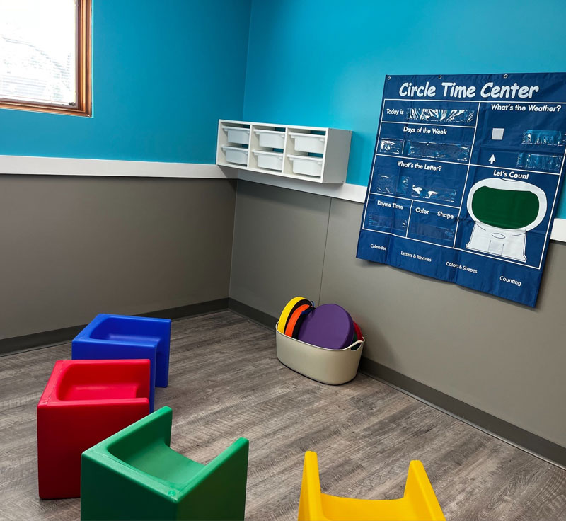 A classroom with a circle time chart on the wall, and 4 chairs in different colors arranged in a semi circle in front of it.