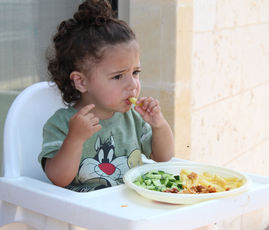 A child in high chair eating a plate of fries, cucumber and meat.