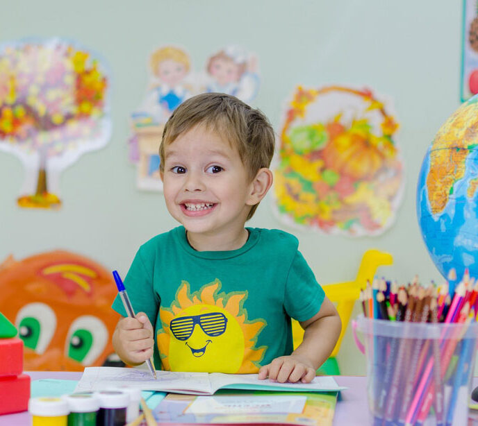 A child smiling in a school room using a pen to draw in a book