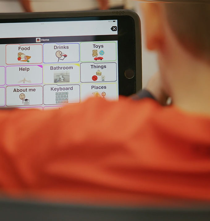 The back of a child wearing a red shirt looking at a tablet with boxes labelled with objects.