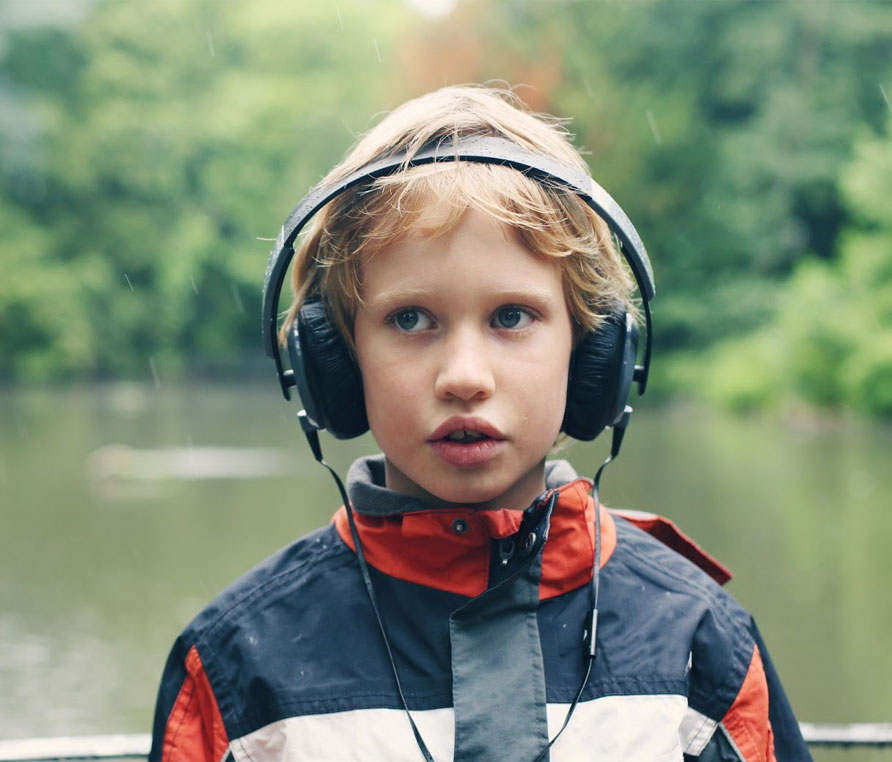 A child stood on a platform in front of a river in the rain wearing a red, white and black jacket with headphones.