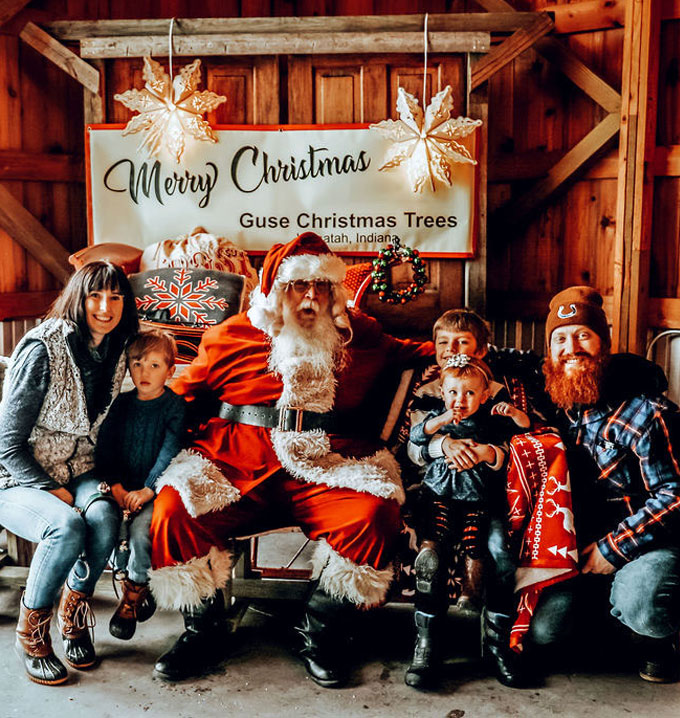 A family with three children all smiling sat around santa claus in front of a white merry christmas sign with hanging paper snowflakes.