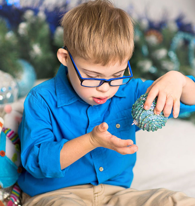 A child wearing a blue shirt and brown trousers sat down in front of holiday decorations and playing with a blue bauble.