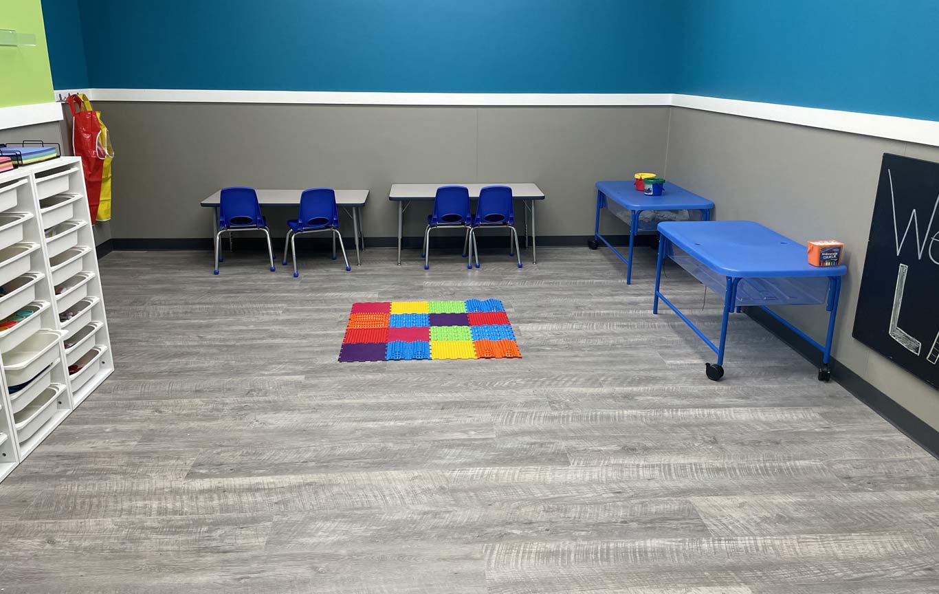 A therapy room with blue and gray walls, a white plastic shelving unit and a multicolored small mat on the gray floor