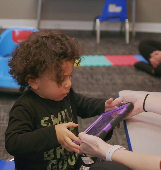 A child with curly hair and wearing a black shirt holding a purple tablet.